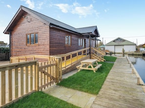 A wooden cabin with a deck, a picnic table, and a small dock extending into a body of water. - A water’s edge setting in the heart of the Norfolk Broads National Park. (Brundall)