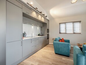 A modern kitchen with grey cabinets, a countertop, and a built-in oven. There is a teal sofa and a small table with a remote control. - A water’s edge setting in the heart of the Norfolk Broads National Park. (Brundall)
