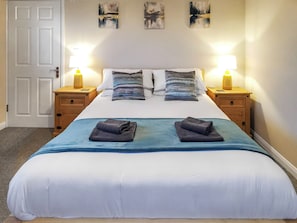 A neatly made bed with a blue and white bedspread, two pillows, two bedside lamps, and a wooden nightstand with a lamp. - A water’s edge setting in the heart of the Norfolk Broads National Park. (Brundall)