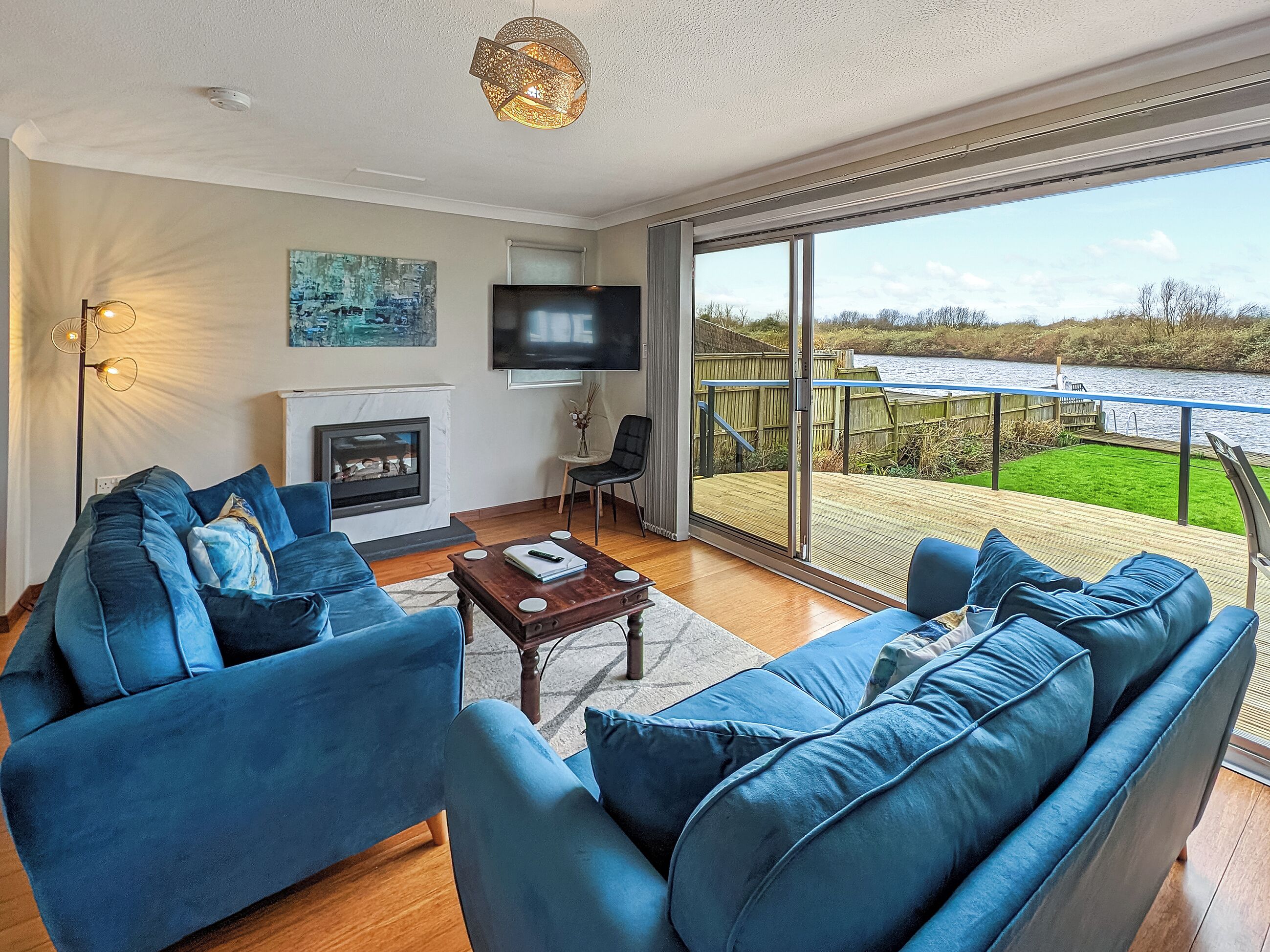 A living room with a large sofa set, a fireplace, a flat-screen TV, and a balcony with a view of a river.