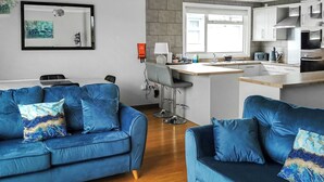 Un saló modern amb dos sofàs blaus, una taula de centre de fusta, i una zona de cuina amb barra i electrodomèstics moderns.