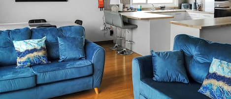 A modern living room with two blue sofas, a wooden coffee table, and a kitchen area with a bar counter and modern appliances.