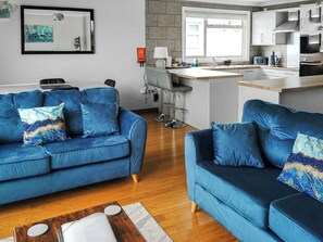 Ein modernes Wohnzimmer mit zwei blauen Sofas, einem Holztisch und einer Küchenzeile mit Barbereich und modernen Geräten.