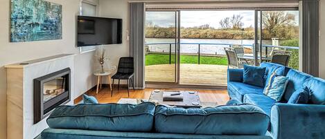 A modern living room with a fireplace, a large sofa, a television, and a view of a deck and water outside.