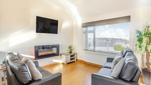 A modern living room with a fireplace, two sofas, a TV, and a large window with blinds. - Math Cottage- UKC6125 (Pentraeth)