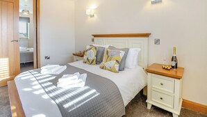 A neatly made bed with a quilt, pillows, and a bottle of champagne on a bedside table. - Math Cottage- UKC6125 (Pentraeth)