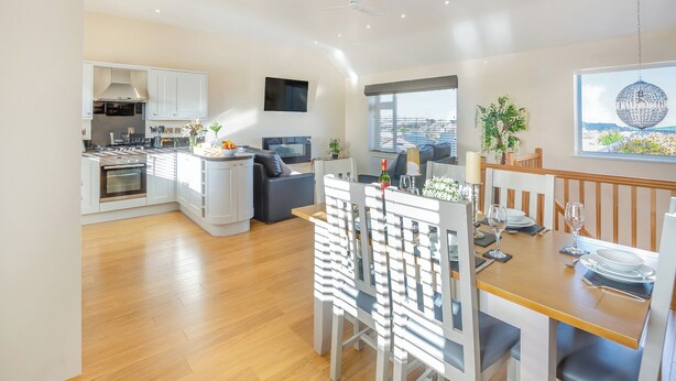 A modern kitchen with white cabinets, a built-in oven, and a dining area with a wooden table and white chairs. - Math Cottage- UKC6125 (Pentraeth)