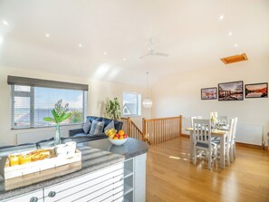A modern kitchen with a breakfast bar, a dining area with a wooden table and chairs, and a ceiling fan. - Math Cottage- UKC6125 (Pentraeth)