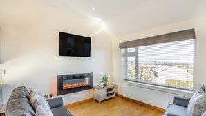 A living room with a fireplace, a flat-screen TV, and a large window with blinds. - Manawydan Cottage - UKC6124 (Pentraeth)