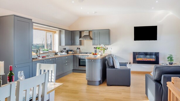 A modern kitchen with grey cabinets, a central island, and a built-in oven. There is a dining area with a white table and chairs, and a living room with a grey sofa and a fireplace. - Manawydan Cottage - UKC6124 (Pentraeth)