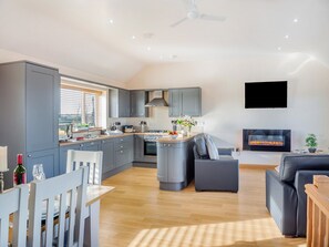 Een moderne keuken met grijze kasten, een centraal eiland en een inbouwoven. Er is een eethoek met een witte tafel en stoelen, en een woonkamer met een grijze bank en een open haard.