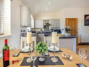 A modern kitchen with white cabinets, a central island with a sink, and a dining area set for two with wine glasses and a bottle. - Pwyll Cottage - UKC6123 (Pentraeth)