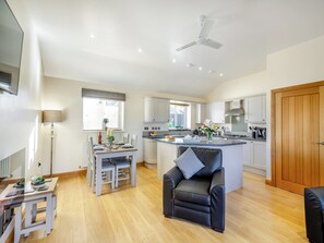 A modern kitchen with white cabinets, a central island, and a dining area with a table and chairs. - Pwyll Cottage - UKC6123 (Pentraeth)
