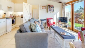 Uma sala de estar moderna com um sofá, uma mesa de jantar e uma televisão. Há uma área de cozinha com armários brancos e uma máquina de lavar louça.