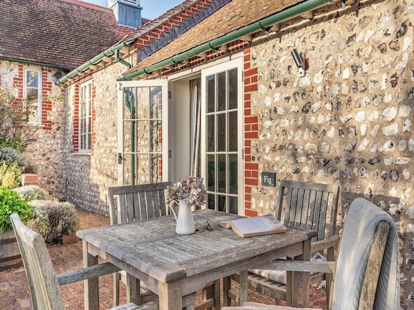 Une maison de campagne en pierre, avec une table et des chaises en bois, une plante en pot et un livre posé sur la table.