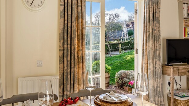Uma área de refeições com uma mesa posta para dois, um relógio na parede e vista para um jardim através de uma porta aberta.