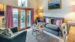 Ein gemütliches Wohnzimmer mit einer Couch, einem runden Couchtisch und einem Esstisch mit Stühlen. Ein Fenster mit Vorhängen, eine Lampe und ein Bild sind an der Wand zu sehen.