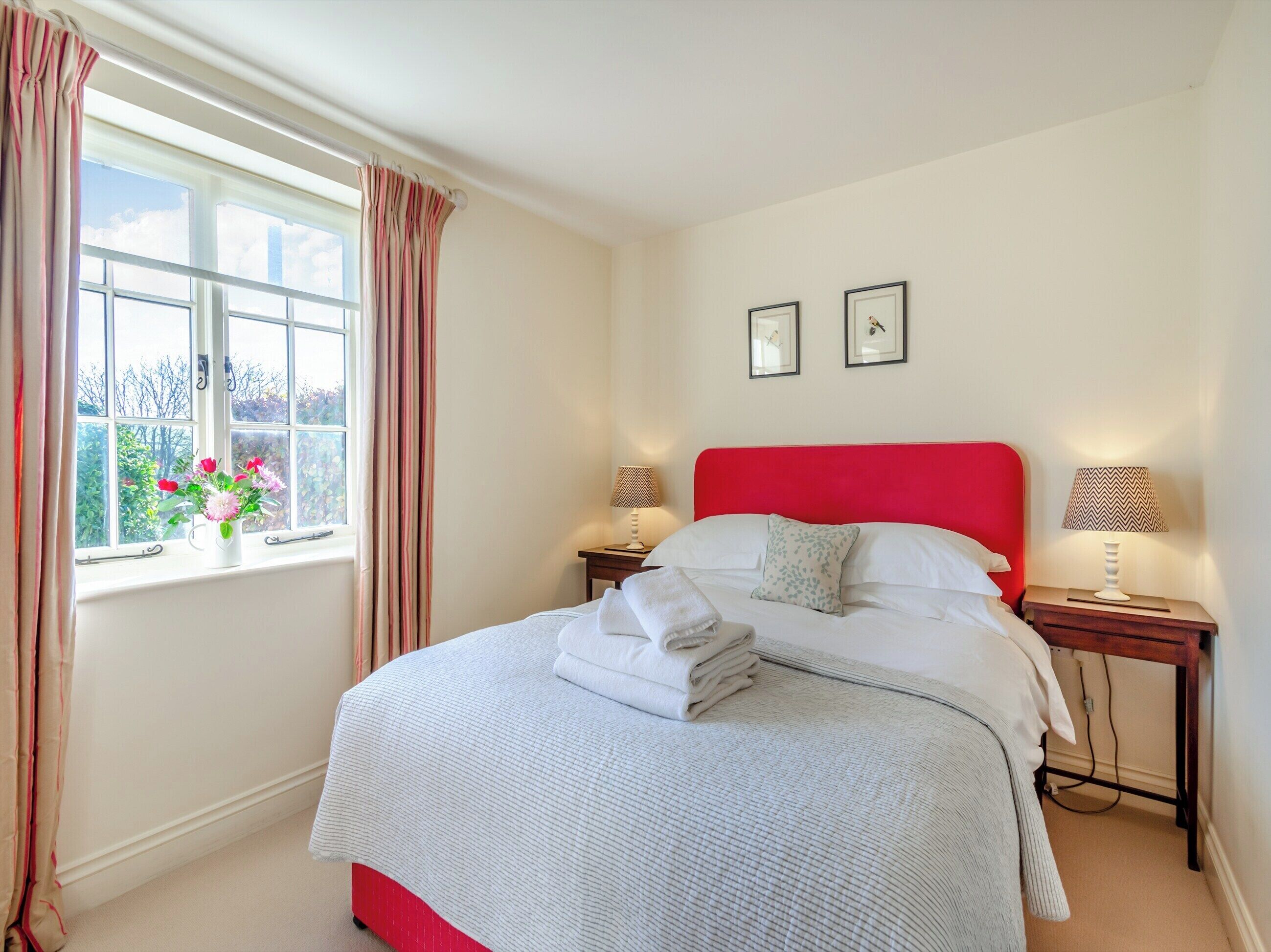 A neatly made bed with a red headboard, two bedside tables with lamps, a window with curtains, and a view of greenery outside.