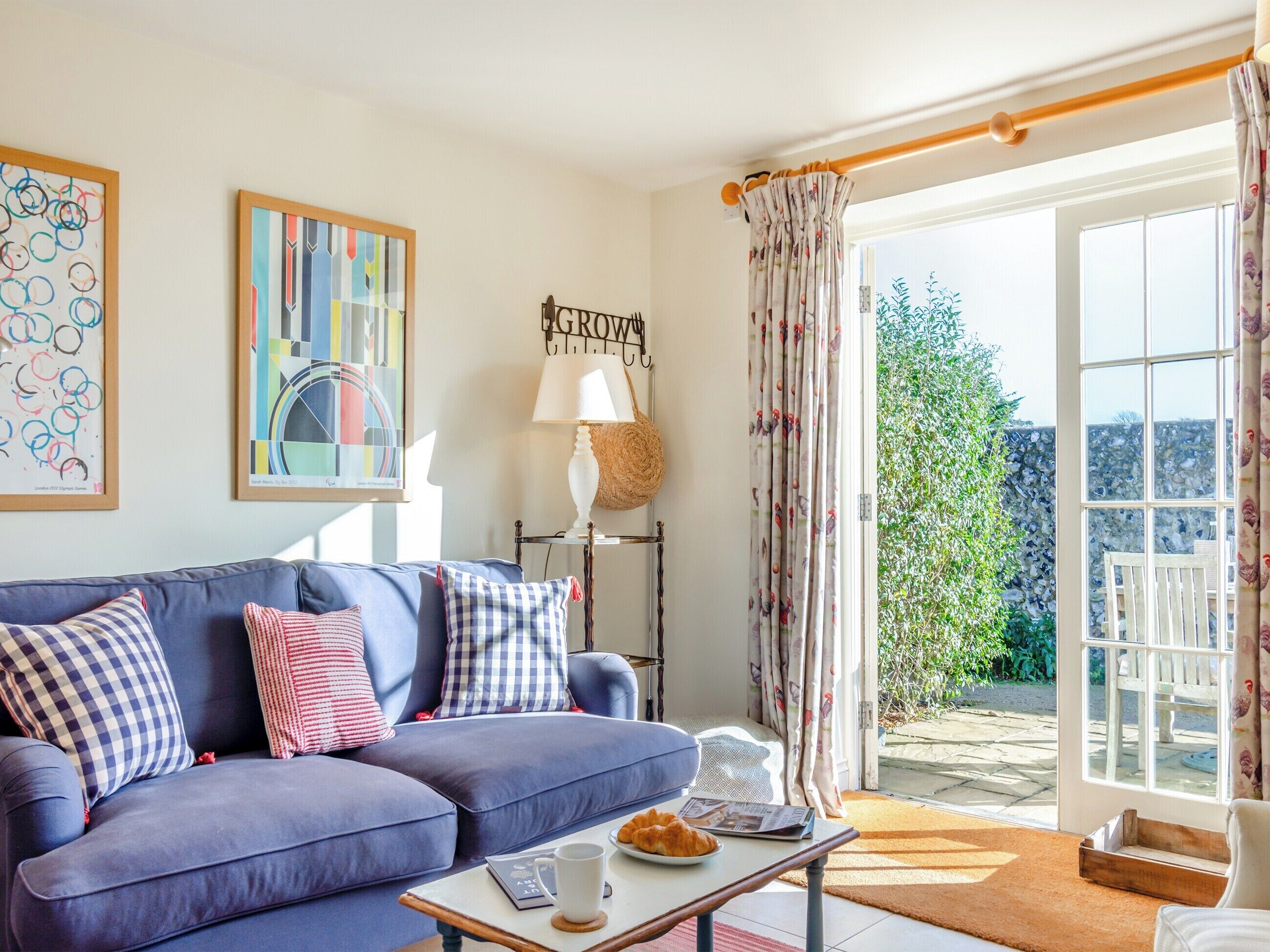Un salon avec un canapé bleu, une table basse sur laquelle se trouve un croissant, une lampe et une vue sur un jardin, visible à travers une porte vitrée.