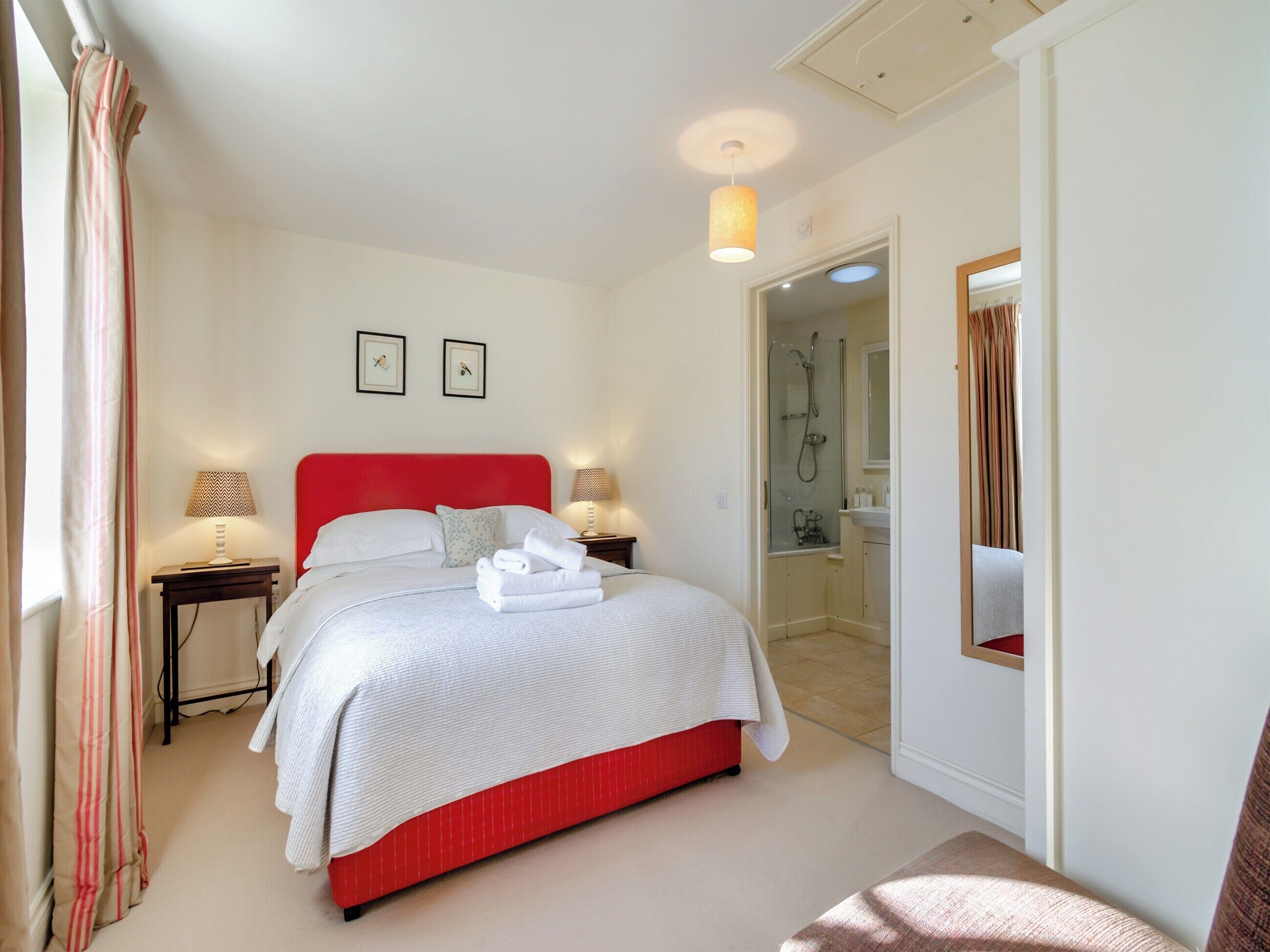 A neatly arranged bedroom with a red headboard, a bed with white linens, two bedside lamps, and a small bedside table.