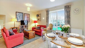 Ein Wohnzimmer mit rotem Sofa, einem Holztisch und einem Esstisch für vier Personen. Ein Fernseher auf einem Standfuß, ein Bücherregal und ein großes Fenster mit Vorhängen sind ebenfalls zu sehen.