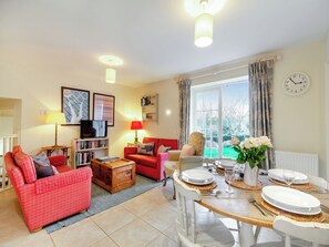 A living room with a red sofa, a wooden coffee table, and a dining table set for four. There is a television on a stand, a bookshelf, and a large window with curtains. - Folly Cottage - UKC6130 (East Dean)