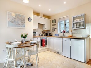 A modern kitchen with white cabinetry, a dining table with chairs, and a window with a view of trees. - Folly Cottage - UKC6130 (East Dean)