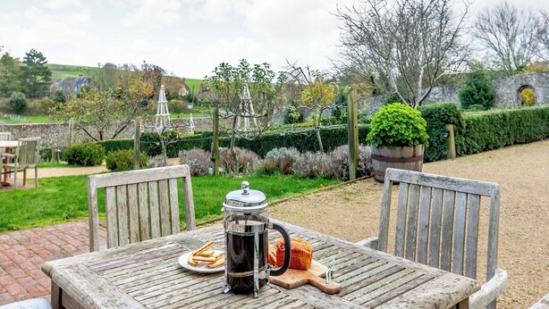 Ein rustikaler Holztisch im Freien mit einer Kaffeekanne, einem Teller mit Keksen und einem Brotlaib, vor einer Gartenkulisse mit Steinmauer und Bäumen.