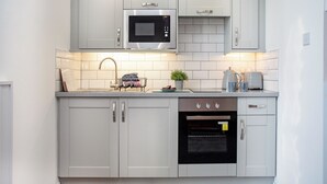 A modern kitchen with white cabinetry, a stainless steel microwave, a built-in oven, and a sink with a dish rack. - Mermaids View - UKC6166 (Paignton)