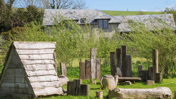 A rustic wooden structure with a sloped roof, surrounded by wooden logs and posts, set against a backdrop of a stone building and a grassy field. - Shepherd's Cottage-UKC6155 (Holbeton)
