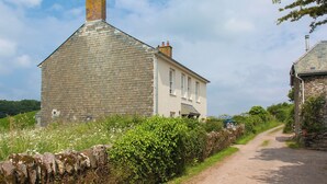 A stone building with a sloped roof and a chimney, adjacent to a white building with a gabled roof and a visible door. - Shepherd's Cottage-UKC6155 (Holbeton)