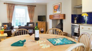 A dining table with wine glasses, a bottle of wine, and a Scrabble game in a room with a fireplace, television, and kitchen area. - Shepherd's Cottage-UKC6155 (Holbeton)