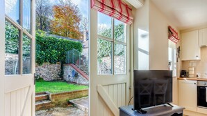 Eine gemütliche Küche mit Fernseher auf einem Schrank, ein Steinweg, der nach draußen führt, und Blick auf einen Garten mit Steinmauer und einer Treppe.