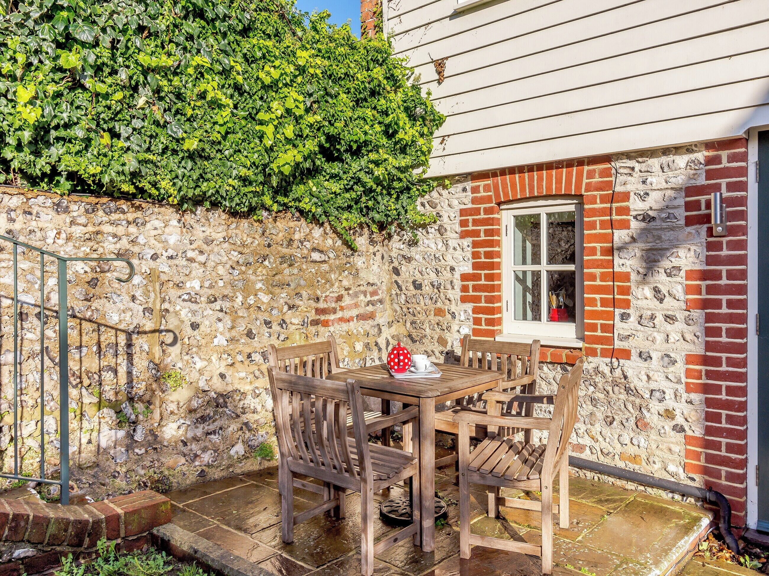 A stone wall with a wooden table and chairs, a window, and a hedge.