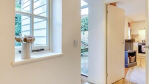 Ein Zimmer mit einem großen Fenster, einer Tür zu einer Küche und einem kleinen Tisch mit einer Vase getrockneter Blumen.
