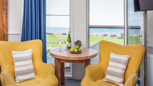 A room with two yellow armchairs, a wooden table with a bottle and glasses, and a view of the sea through large windows. - The Little Lobster - UKC6167 (Paignton)