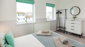 Un dormitorio con una cama, dos ventanas con cortinas verdes, un radiador, un perchero negro, un tocador blanco y un espejo redondo.