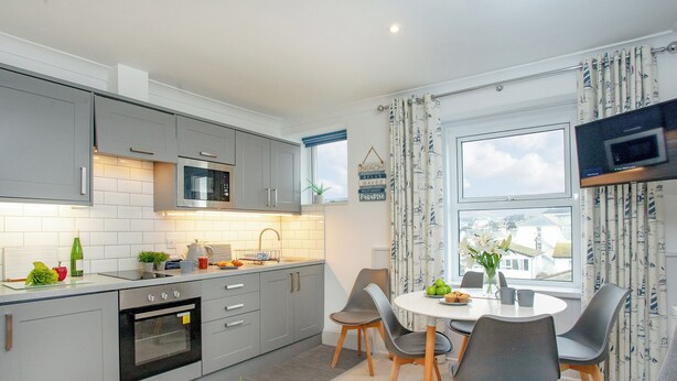 Een moderne keuken met grijze kastjes, een magnetron en een spoelbak. Er is een zithoek met een tafel en stoelen, en een tv aan de wand bevestigd.