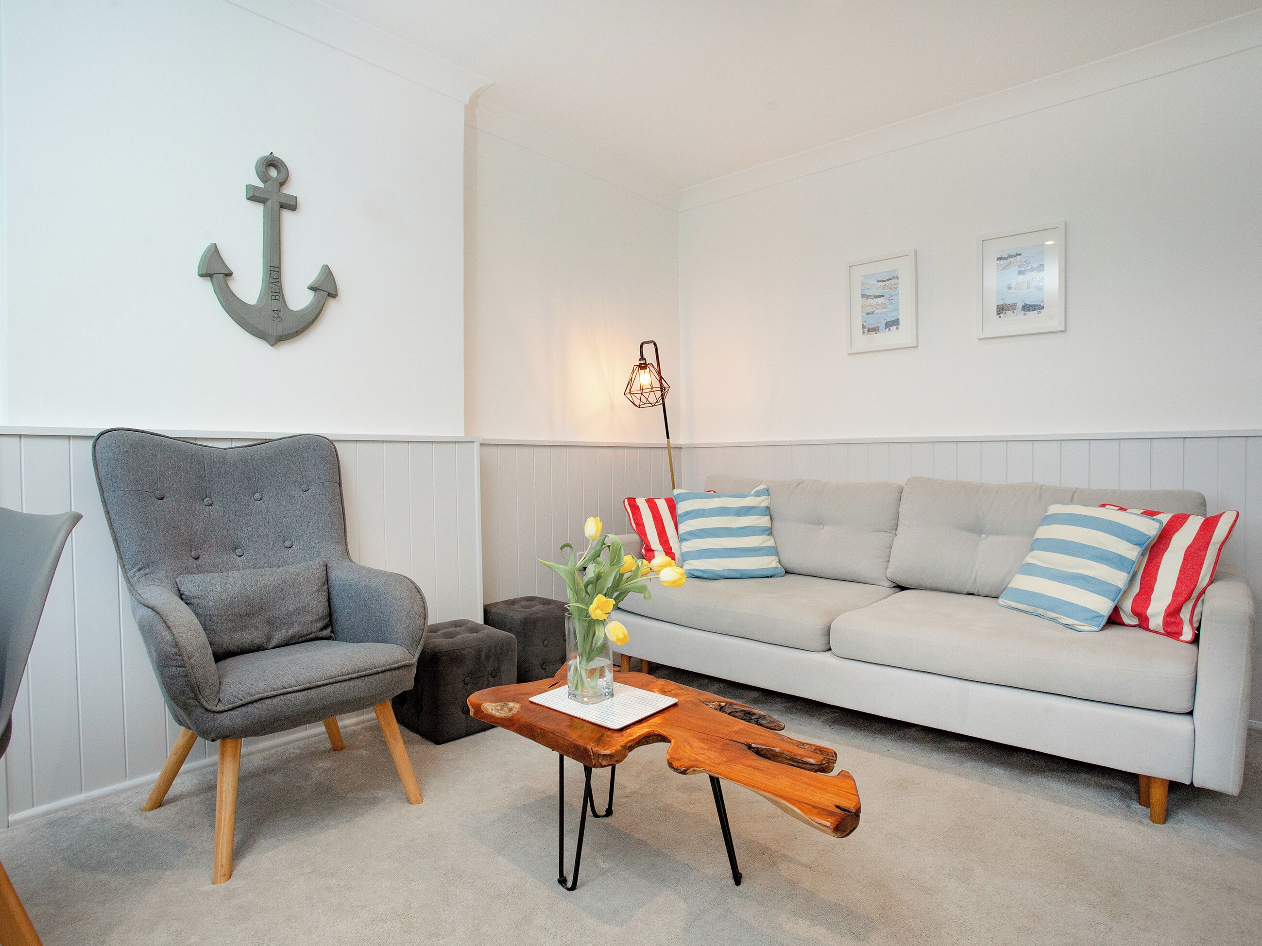 Uma sala de estar com uma poltrona cinza, um sofá branco com almofadas listradas, uma mesa de centro de madeira e um enfeite de âncora fixado na parede.