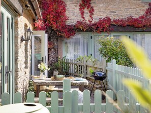 A stone building with a wooden deck, a table with outdoor seating, a grill, and a white picket fence. - Daisy Cottage - UKC6177 (Marldon, near Paignton)