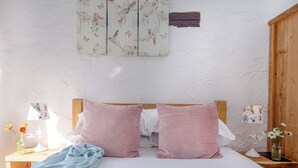 Een slaapkamer met een houten hoofdbord, twee roze kussens, een bloemenbedekking, een nachtkastje met een lamp en een kunstwerk aan de muur.