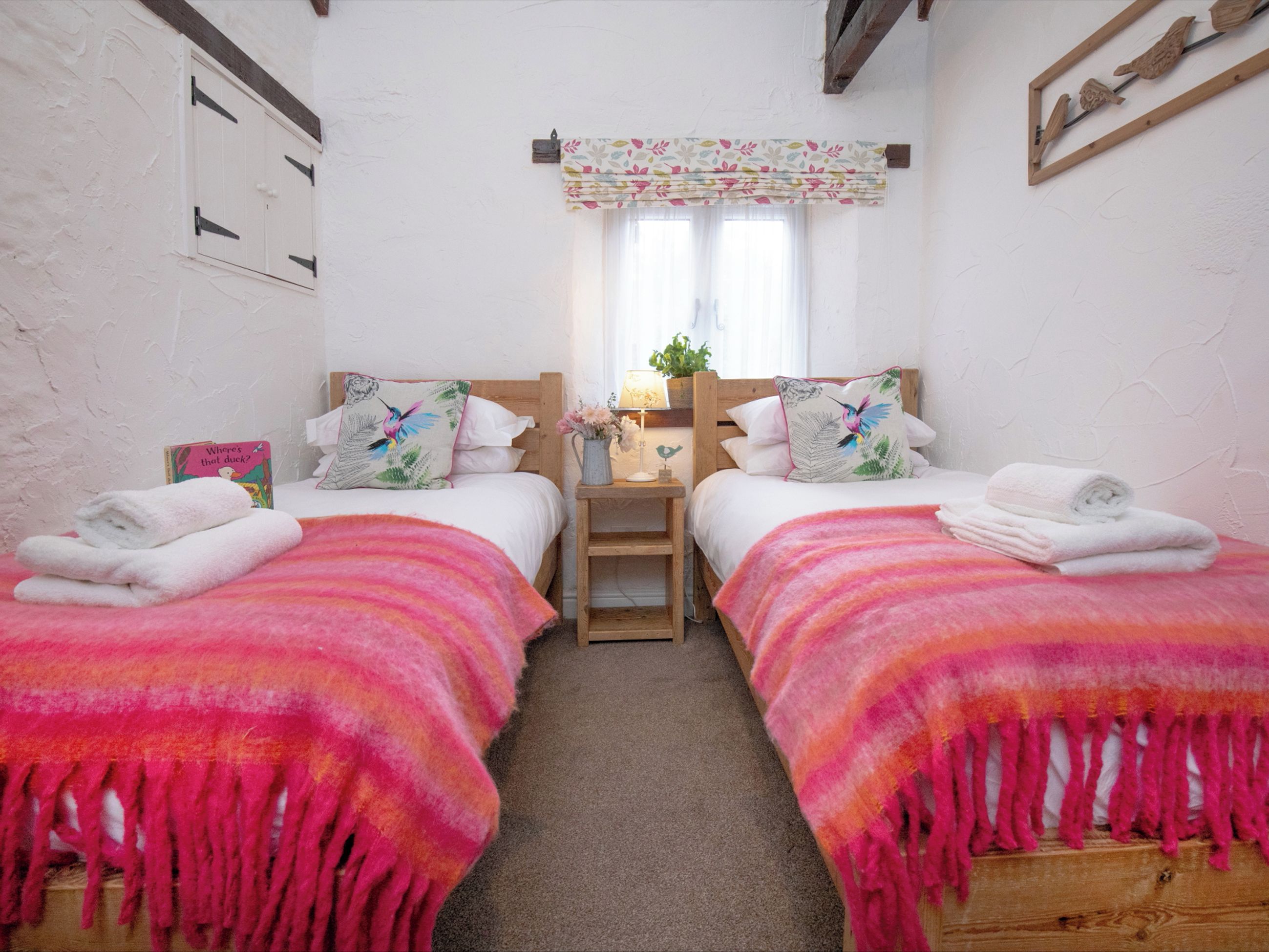 A room with two beds, each with a pink and orange blanket, a small bedside table with a lamp, and a window with a floral curtain.