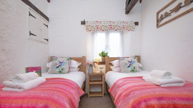 Een kamer met twee bedden, elk met een roze en oranje plaid, een klein nachtkastje met een lamp en een raam met een bloemenprint gordijn.