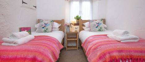 A room with two beds, each with a pink and orange blanket, a small bedside table with a lamp, and a window with a floral curtain.