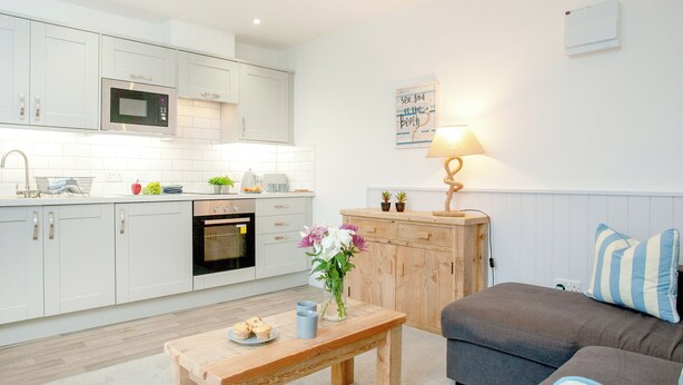 A modern kitchen with white cabinets, a microwave, and an oven. There is a wooden table with a vase of flowers and a plate of muffins. A gray sofa and striped pillows are in the living area. - Anchors Away - UKC6170 (Paignton)