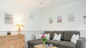 A living room with a grey sofa, wooden side table, and a lamp. There are wall-mounted pictures and a sign on the wall. - Anchors Away - UKC6170 (Paignton)