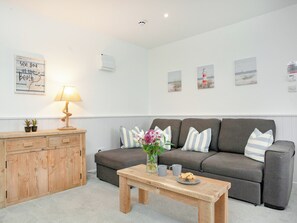 Ein Wohnzimmer mit grauem Sofa, Holzbeistelltisch und einer Lampe. An den Wänden hängen Bilder und es gibt ein Schild.