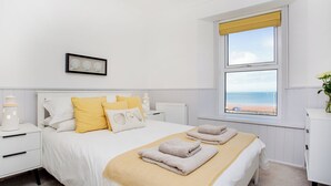 Un dormitori amb un llit gran, una tauleta de nit, una meseta al costat del llit, una finestra amb vista a la platja i una fotografia emmarcada a la paret.