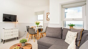 Un saló modern amb un sofà gris, una taula de centre rodona de fusta amb fruita, una zona de menjador amb taula i cadires blanques, un armari blanc i un televisor penjat a la paret.