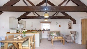 A rustic interior with exposed wooden beams, a dining area with a wooden table and chairs, a kitchen with a white countertop, a living room with a sofa and a coffee table, and a fireplace. - Jasmine Cottage - UKC6179 (Marldon, near Paignton)